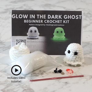 May include: A glow in the dark ghost beginner crochet kit featuring a white yarn ball, a bag of stuffing, a red crochet hook, black safety eyes, stitch markers, and a finished white crocheted ghost with black eyes and a printed instruction card.