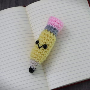 May include: A crocheted pencil with a pink eraser, yellow body, and gray band. The pencil has a smiling face with black eyes and a black mouth. The pencil is lying on a lined notebook.
