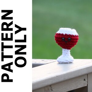 May include: A crocheted wine glass with a red wine filling and white stem. The glass has a cute face with black eyes.