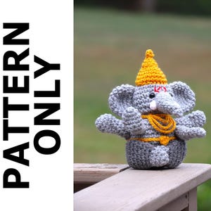 May include: Crocheted gray elephant with a yellow hat and a yellow beaded necklace. The elephant is sitting on a wooden surface.