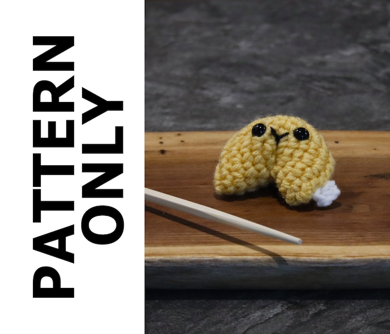 May include: A crocheted yellow fortune cookie with black eyes and a white accent, resting on a wooden surface. A pair of wooden chopsticks is placed diagonally. The text "PATTERN ONLY" is printed vertically on the left side.