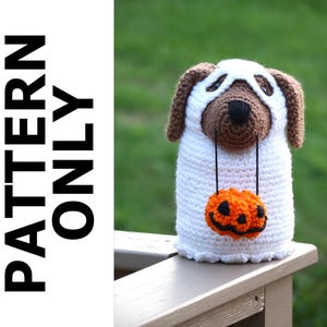 May include: A crocheted ghost dog wearing a white ghost costume with a brown face and a small orange pumpkin hanging from its neck. The dog is sitting on a wooden surface.