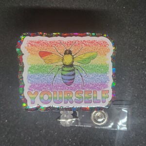 Puede incluir: Un portagafete con brillo de colores arcoíris. El portagafete presenta una abeja con un fondo de colores arcoíris y el texto "YOURSELF".