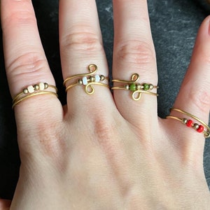 Könnte beinhalten: Eine Hand trägt fünf goldene Drahtringe mit unterschiedlich farbigen Perlen. Die Ringe sind auf dem Mittelfinger übereinander gelegt. Die Perlen sind rot, weiß, grün und braun.
