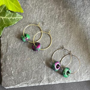 Könnte beinhalten: Zwei Paar goldene Creolen. Jeder Ohrring hat eine farbenfrohe, runde Perle mit grünen und magentafarbenen Farbtönen. Die Ohrringe werden auf einer grauen, strukturierten Oberfläche mit grünen Blättern und einem Stück Treibholz im Hintergrund präsentiert.