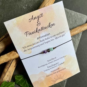 Könnte beinhalten: Ein schwarzes Armband mit Amethyst-, grünen und klaren Perlen, präsentiert mit einer Karte. Die Karte enthält deutschen Text, darunter "Angst & Panikattacken" und eine Affirmation. Der Hintergrund umfasst Holz und einen dunkelgrauen Stein.