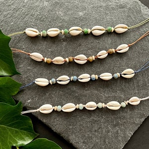 Könnte beinhalten: Vier Muschel- und Perlenarmbänder mit unterschiedlich farbigen Perlen. Die Armbänder liegen auf einer grauen Steinoberfläche mit grünen Blättern in der linken unteren Ecke.