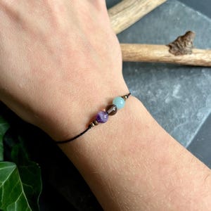 Puede incluir: Una pulsera de hilo negro con tres cuentas de piedras preciosas: una amatista morada, un cuarzo ahumado marr&oacute;n y una aguamarina azul claro. La pulsera tiene peque&ntilde;os detalles met&aacute;licos dorados. La pulsera se muestra en una mu&ntilde;eca.