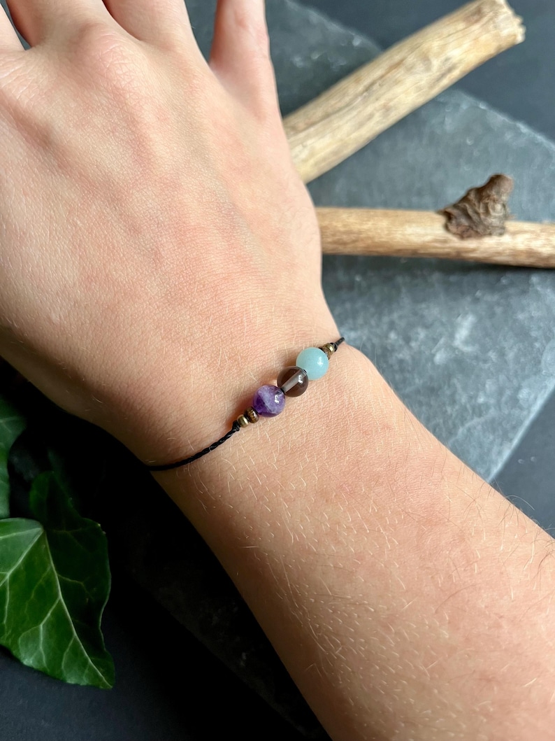 Puede incluir: Una pulsera de hilo negro con tres cuentas de piedras preciosas: una amatista morada, un cuarzo ahumado marr&oacute;n y una aguamarina azul claro. La pulsera tiene peque&ntilde;os detalles met&aacute;licos dorados. La pulsera se muestra en una mu&ntilde;eca.