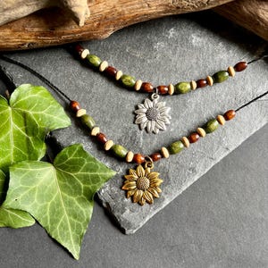 Puede incluir: Dos collares con cuentas de madera en tonos marrones y verdes. Cada collar presenta un dije de girasol, uno plateado y otro dorado.