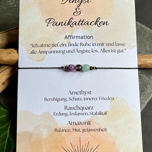 Puede incluir: Un brazalete de cord&oacute;n negro con tres cuentas de piedras preciosas: amatista, cuarzo ahumado y amazonita. El brazalete se muestra en una tarjeta con texto en alem&aacute;n sobre la ansiedad y los ataques de p&aacute;nico.