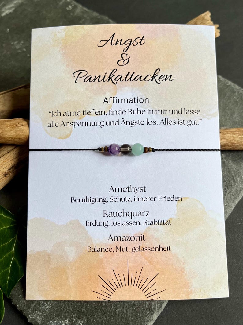 Puede incluir: Un brazalete de cord&oacute;n negro con tres cuentas de piedras preciosas: amatista, cuarzo ahumado y amazonita. El brazalete se muestra en una tarjeta con texto en alem&aacute;n sobre la ansiedad y los ataques de p&aacute;nico.