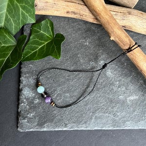 Puede incluir: Una pulsera de cord&oacute;n negro con tres cuentas: una cuenta azul claro, una cuenta morada y una cuenta dorada. La pulsera se muestra sobre una superficie de piedra gris oscuro, con hojas verdes y palos de madera en el fondo.