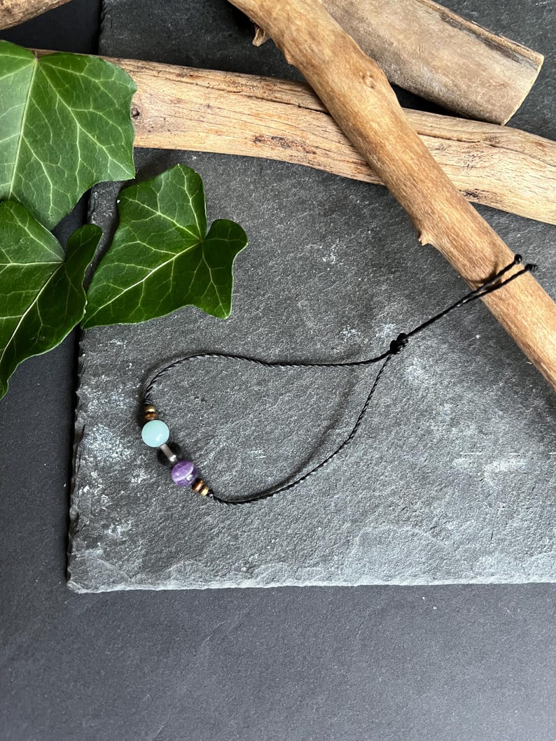 Puede incluir: Una pulsera de cord&oacute;n negro con tres cuentas: una cuenta azul claro, una cuenta morada y una cuenta dorada. La pulsera se muestra sobre una superficie de piedra gris oscuro, con hojas verdes y palos de madera en el fondo.