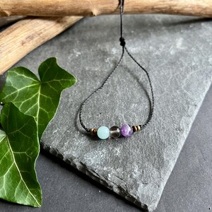 Puede incluir: Una pulsera de cord&oacute;n negro con tres cuentas de piedras preciosas: una azul claro, una gris ahumado y una piedra p&uacute;rpura. La pulsera tiene peque&ntilde;as cuentas doradas a cada lado de las piedras preciosas. La pulsera se exhibe sobre una superficie de piedra gris.