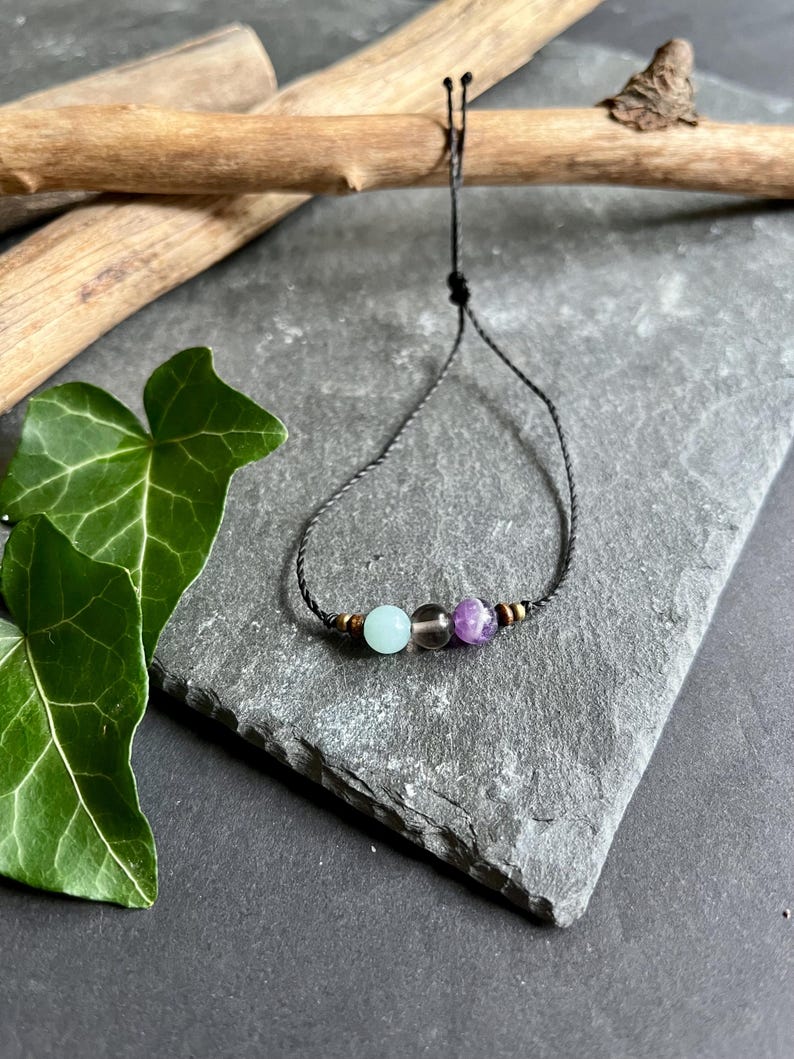 Puede incluir: Una pulsera de cord&oacute;n negro con tres cuentas de piedras preciosas: una azul claro, una gris ahumado y una piedra p&uacute;rpura. La pulsera tiene peque&ntilde;as cuentas doradas a cada lado de las piedras preciosas. La pulsera se exhibe sobre una superficie de piedra gris.