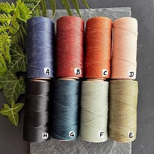 Puede incluir: Un surtido de ocho carretes de hilo encerado en varios colores, incluyendo azul, rojo, naranja y beige. Los carretes est&aacute;n etiquetados de la A a la H. Los hilos est&aacute;n dispuestos sobre una superficie gris, con follaje verde en el fondo.