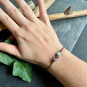 Puede incluir: Un brazalete de hilo negro con tres cuentas de piedras preciosas en tonos morado, marr&oacute;n y verde azulado. El brazalete se muestra en una mu&ntilde;eca sobre un fondo oscuro con elementos de madera y hojas verdes.