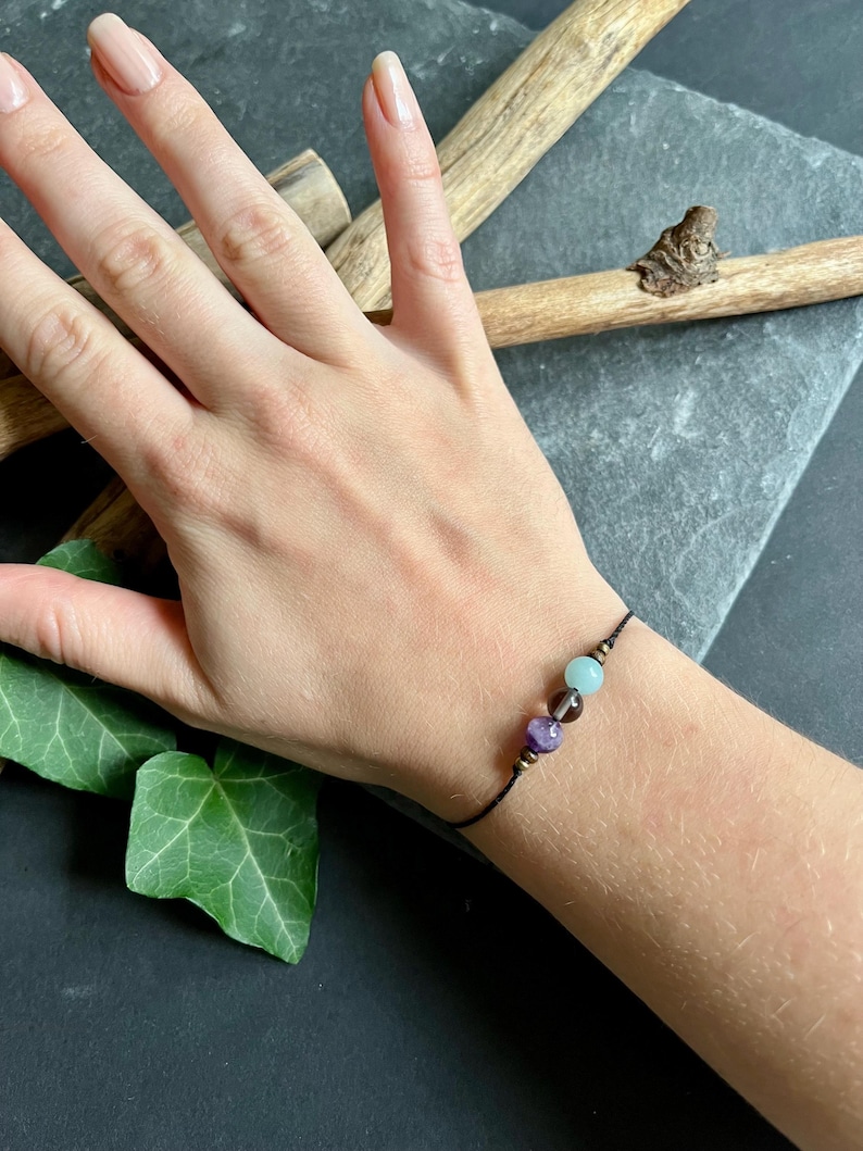 Puede incluir: Un brazalete de hilo negro con tres cuentas de piedras preciosas en tonos morado, marr&oacute;n y verde azulado. El brazalete se muestra en una mu&ntilde;eca sobre un fondo oscuro con elementos de madera y hojas verdes.