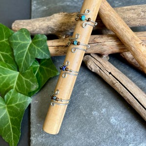 Könnte beinhalten: Handgefertigte Drahtringe mit farbigen Perlen, präsentiert auf einem Holzstab. Die Ringe zeigen Perlen in Blau-, Braun- und Türkistönen. Der Stab liegt vor einem Hintergrund aus natürlichen Elementen, darunter Holzstücke und grünes Efeu.