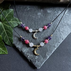 Puede incluir: Dos collares con cuentas de piedras preciosas en tonos morados, rosas y blancos. Cada collar presenta un colgante de media luna. Los collares se exhiben sobre una superficie oscura y texturizada.