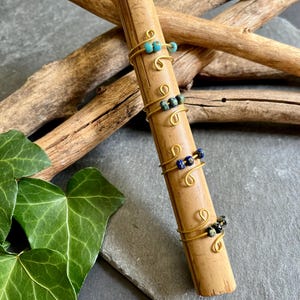 Puede incluir: Anillos de alambre hechos a mano con cuentas de colores. Los anillos están envueltos alrededor de una clavija de madera, mostrando su diseño. Las cuentas son en tonos turquesa, azul y negro. Se exhiben sobre una superficie gris oscura.
