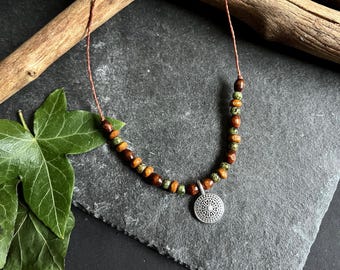Può includere: Una collana fatta a mano con un ciondolo d'argento dal design intricato. La collana ha un cordino marrone ed è adornata da perline di legno nei toni del marrone, verde e arancione. Presentata su una superficie grigio scuro.