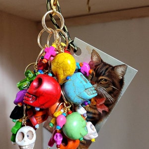 May include: A colorful keychain with a variety of charms, including skulls, beads, and a small tag with a cat image. The charms are made of different materials, including wood, metal, and glass. The keychain is attached to a gold-colored ring.