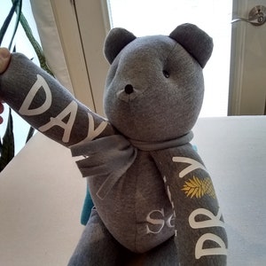 May include: A gray teddy bear with a white scarf and the words "Day Dream" written on its arms. The bear is sitting on a white surface.