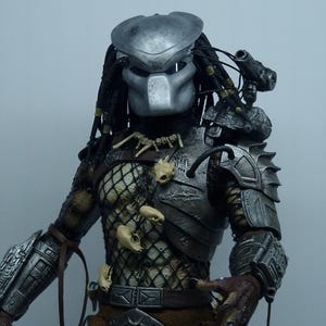 Peut inclure: Figurine détaillée d'un Predator, avec un casque argenté et des dreadlocks, et un bio-masque. La figurine présente une armure complexe, une poitrine en forme de filet, des ornements en forme d'os et une ceinture marron. La figurine est en pose dynamique.