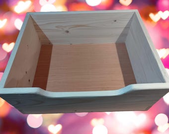 May include: A rectangular wooden box with a curved cutout on one side. The box is made of light-coloured wood with a natural finish. The interior is a lighter shade of wood. The background is a blurred mix of pink, red, and white.
