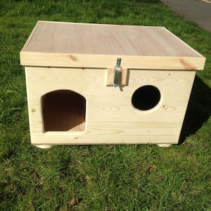 May include: A light-coloured wooden cat house with a rectangular design and a flat roof. It has an arched entrance and a circular opening. A metal latch secures the lid. The house rests on small, rounded feet.