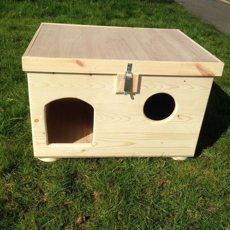 May include: A light-coloured wooden cat house with a rectangular design and a flat roof. It has an arched entrance and a circular opening. A metal latch secures the lid. The house rests on small, rounded feet.