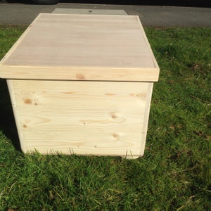 May include: A rectangular wooden storage box made from light-coloured, unfinished wood. The box has a flat lid and is positioned on green grass. The wood grain is visible on the sides and top.