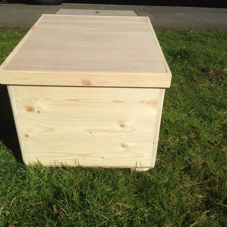 May include: A rectangular wooden storage box made from light-coloured, unfinished wood. The box has a flat lid and is positioned on green grass. The wood grain is visible on the sides and top.