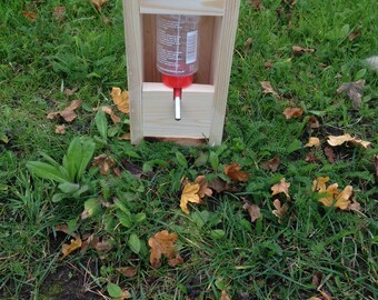 May include: A wooden bird feeder with a clear plastic water bottle and a red spout. The feeder is sitting on a bed of green grass and brown leaves.