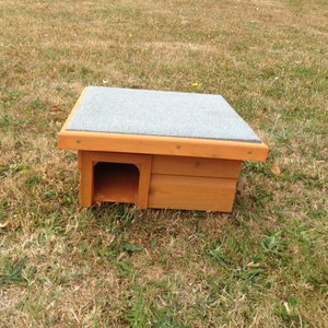 May include: A wooden birdhouse with a grey roof and a small entrance hole. The birdhouse is sitting on a patch of brown grass.
