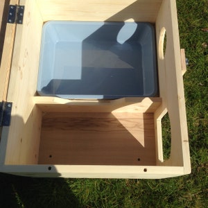 May include: A light-coloured wooden pet house with a grey plastic tray inside. The house has a rectangular shape with a hinged door and a cutout handle on the side. The house is sitting on green grass.
