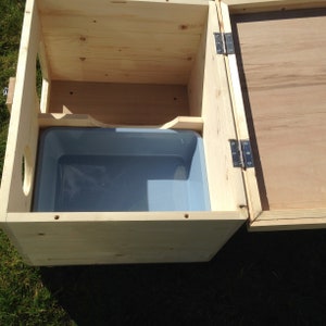 May include: A wooden pet house with a hinged lid and a grey plastic tub inside. The house is made of light-coloured wood and has a circular opening on one side. The lid is open, revealing the interior.