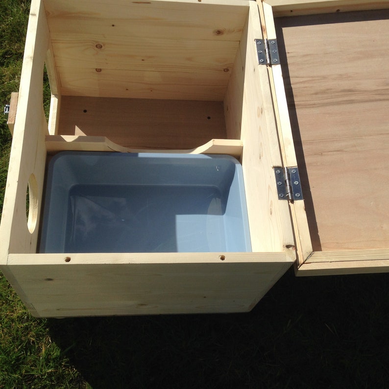 May include: A wooden pet house with a hinged lid and a grey plastic tub inside. The house is made of light-coloured wood and has a circular opening on one side. The lid is open, revealing the interior.