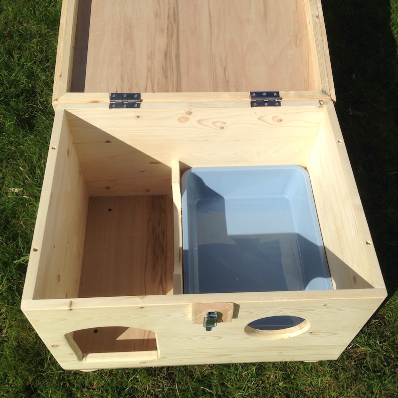 May include: A light-coloured wooden cat house with an open lid. The interior features a light blue litter tray and a separate compartment. The front has two oval openings and a latch. The cat house is on green grass.