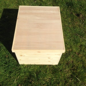 May include: A rectangular wooden box with a light-coloured, flat lid. The box is made of untreated wood, showing the natural grain. It sits on green grass. The box is likely for storage or display, with a simple, clean design.