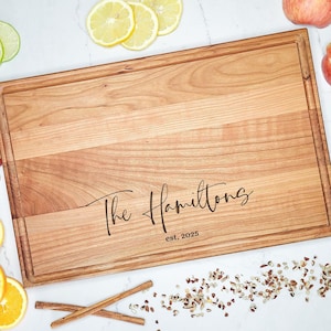 May include: A large, rectangular wooden cutting board with a natural wood finish. The board has a raised edge and is engraved with the text "The Hamiltons est. 2025".