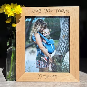 May include: A light-colored wooden picture frame with the handwritten text "I Love You mom" at the top and "Tatum" with a heart at the bottom. The frame holds a photo of a mother and child. A vase of yellow flowers is to the left.