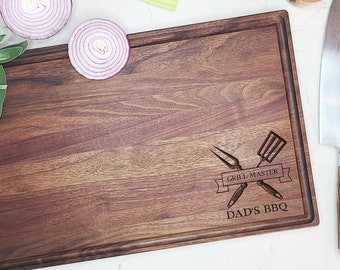 Może przedstawiać: Duża, prostokątna, ciemnobrązowa drewniana deska do krojenia z wzorem mistrza grilla i wygrawerowanym tekstem "Dad's BBQ" na powierzchni. Deska jest otoczona różnymi składnikami, w tym czerwoną cebulą, pomidorami i ziołami.