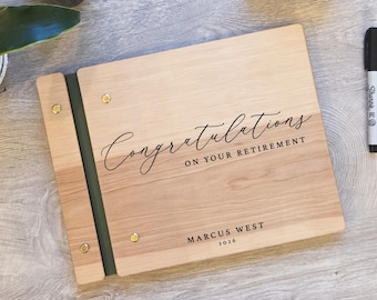 May include: A rectangular wooden photo album with a dark green spine and gold metal binding screws. The cover features the engraved text "Congratulations ON YOUR RETIREMENT" followed by the name "MARCUS WEST" and the year "2026" in black ink.