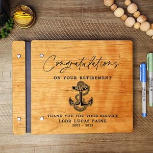 May include: A wooden guest book with a navy blue anchor and the text "Congratulations on your retirement. Thank you for your service. LCDR Lucas Paine 2005 - 2025".