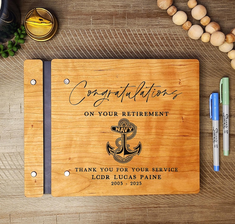 May include: A wooden guest book with a navy blue anchor and the text "Congratulations on your retirement. Thank you for your service. LCDR Lucas Paine 2005 - 2025".