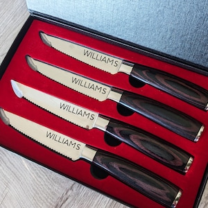 May include: A set of four steak knives in a black gift box with a red interior. Each knife has a serrated blade with the name "WILLIAMS" etched on it, and a dark wood-grain handle.