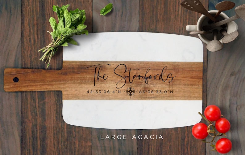 May include: A large acacia wood cutting board with a white marble top. The board is engraved with "The Stanfords" and coordinates, "42&deg;53'06.4"N 83&deg;36'33.0"W".
